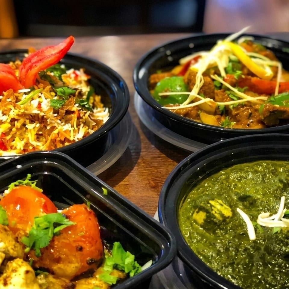 Four black bowls of Indian food, including biryani, curry, palak paneer, and chicken tikka.